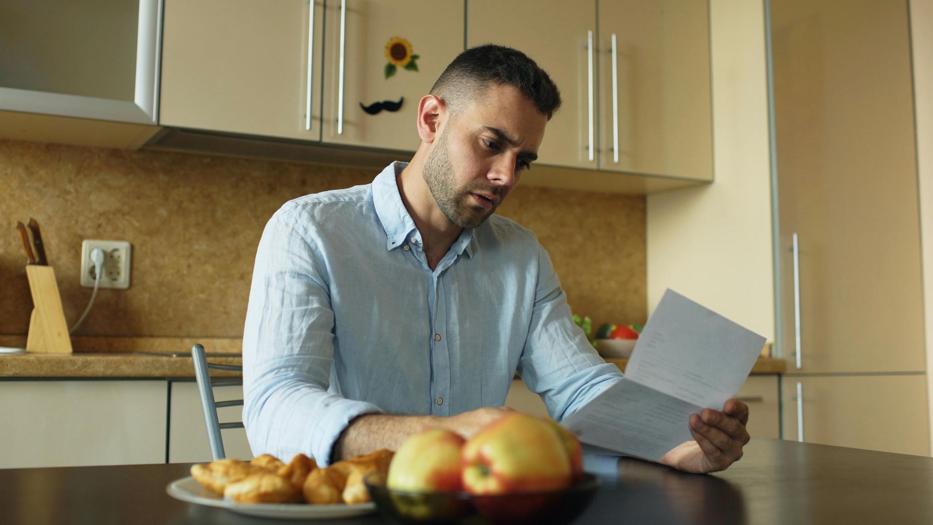 Man in a kitchen reading a document, apples on table.