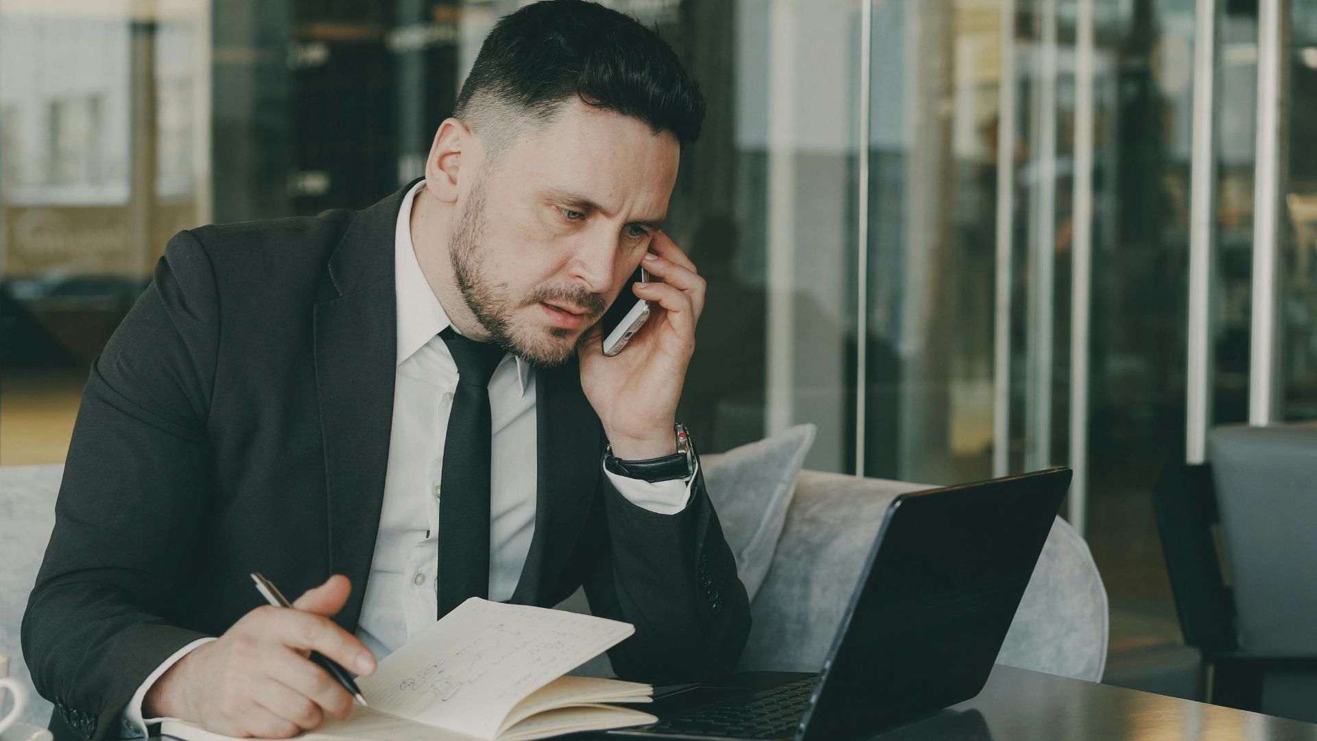Businessman talks on phone while writing notes in a modern cafe.