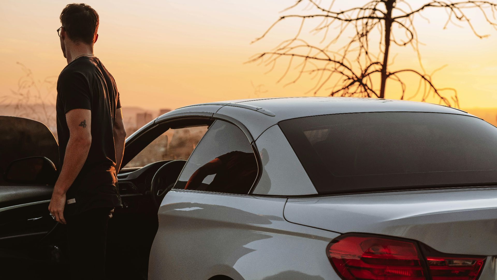A man stands beside his car, enjoying a scenic sunset in an open landscape.