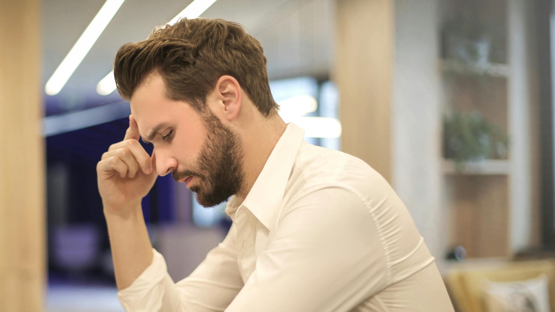 A thoughtful man in a shirt works on his laptop at a modern indoor office space.