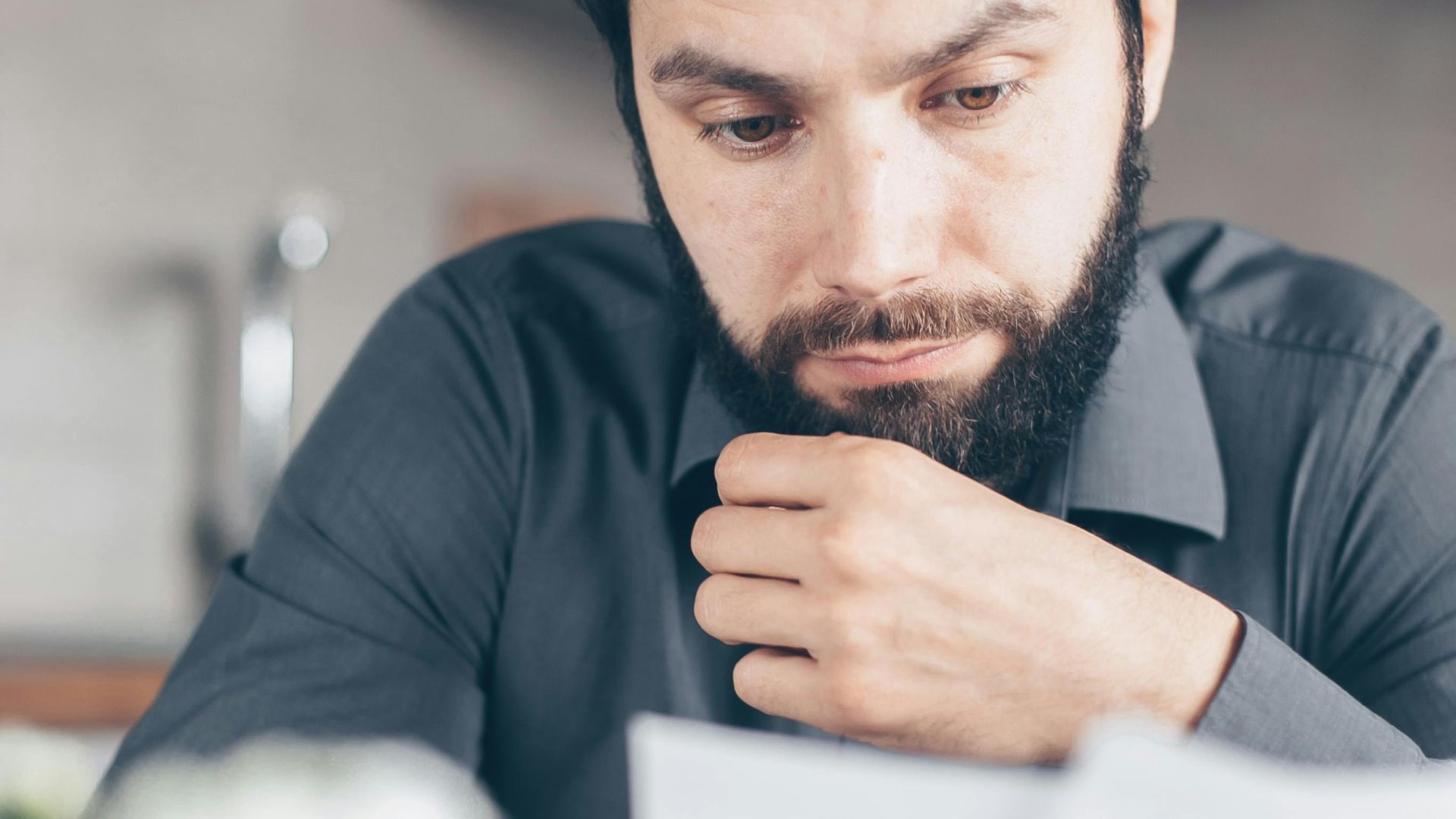 A bearded man in a gray shirt intently reads documents while seated indoors, appearing concerned.