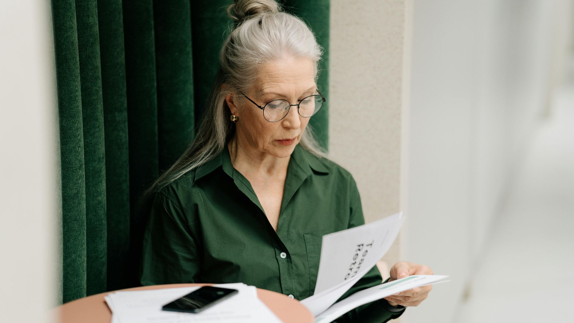 Elderly woman with gray hair reading papers at a small table indoors, focused and thoughtful.