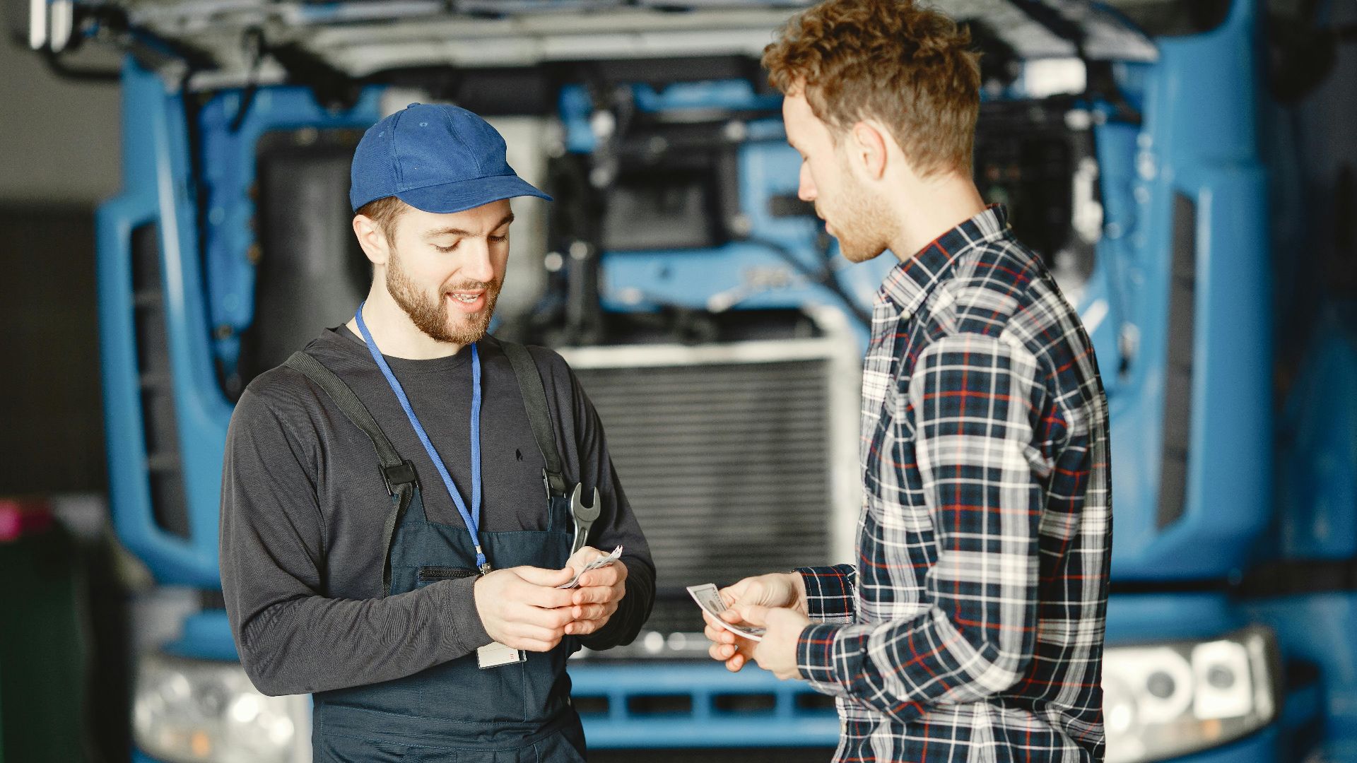 Auto mechanic in blue uniform receives payment from customer in a garage setting with a truck in the background.
