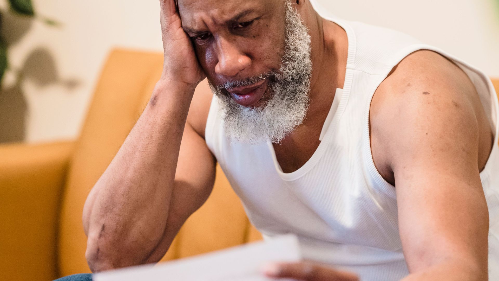 A worried man sitting on a sofa examining documents, expressing anxiety.