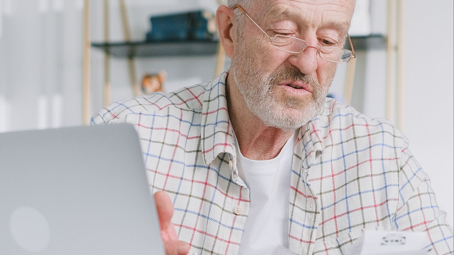 Elderly man reading papers at home with a laptop, emphasizing financial planning.