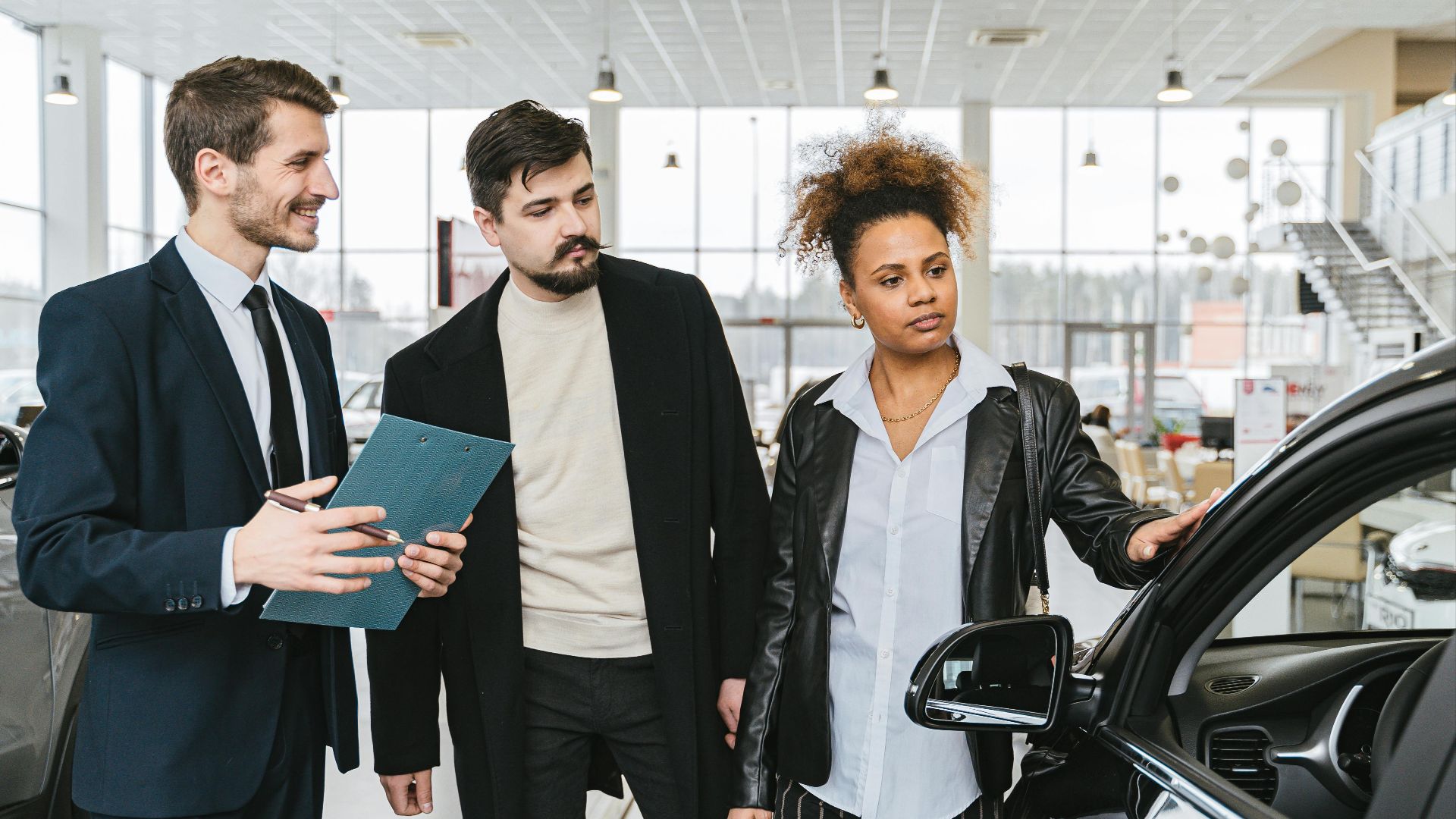 An interracial couple consults with a salesman at a car dealership, exploring vehicle options.