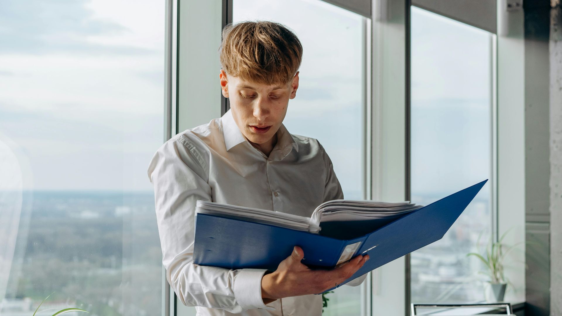 Businessman reading documents in an office with a city view, holding a blue binder.