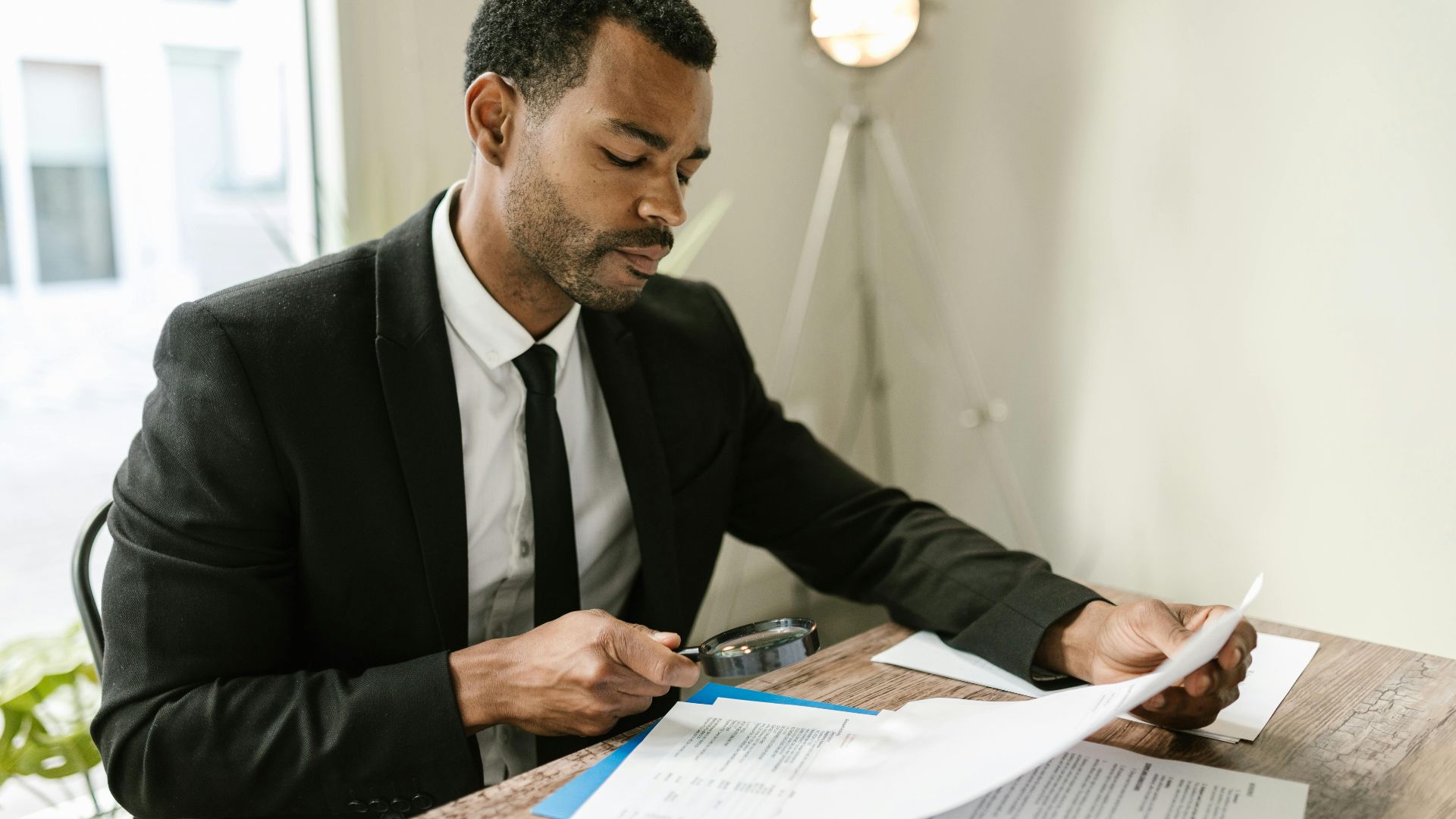 Professional man examines paperwork with magnifying glass at office desk, highlighting attention to detail.