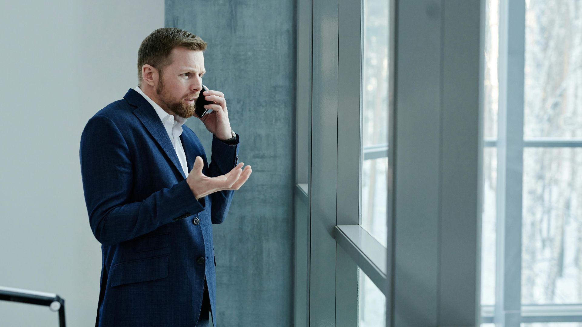Businessman using smartphone while standing indoors by glass window, expressing concern.