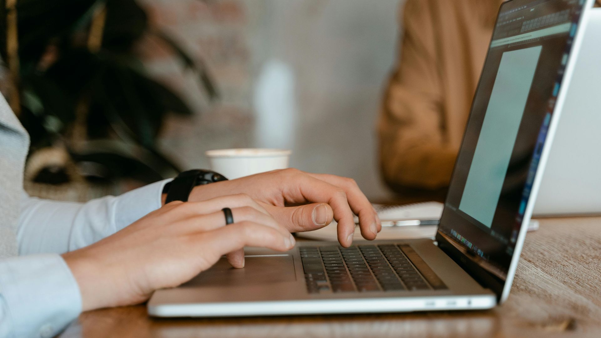 A person typing on a laptop at a wooden table in a modern office setting.