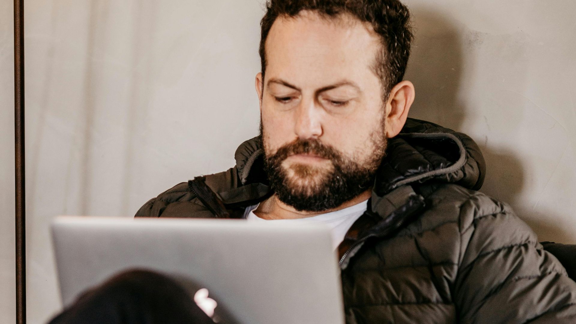 Bearded man focused on typing with a laptop indoors, wearing a jacket.
