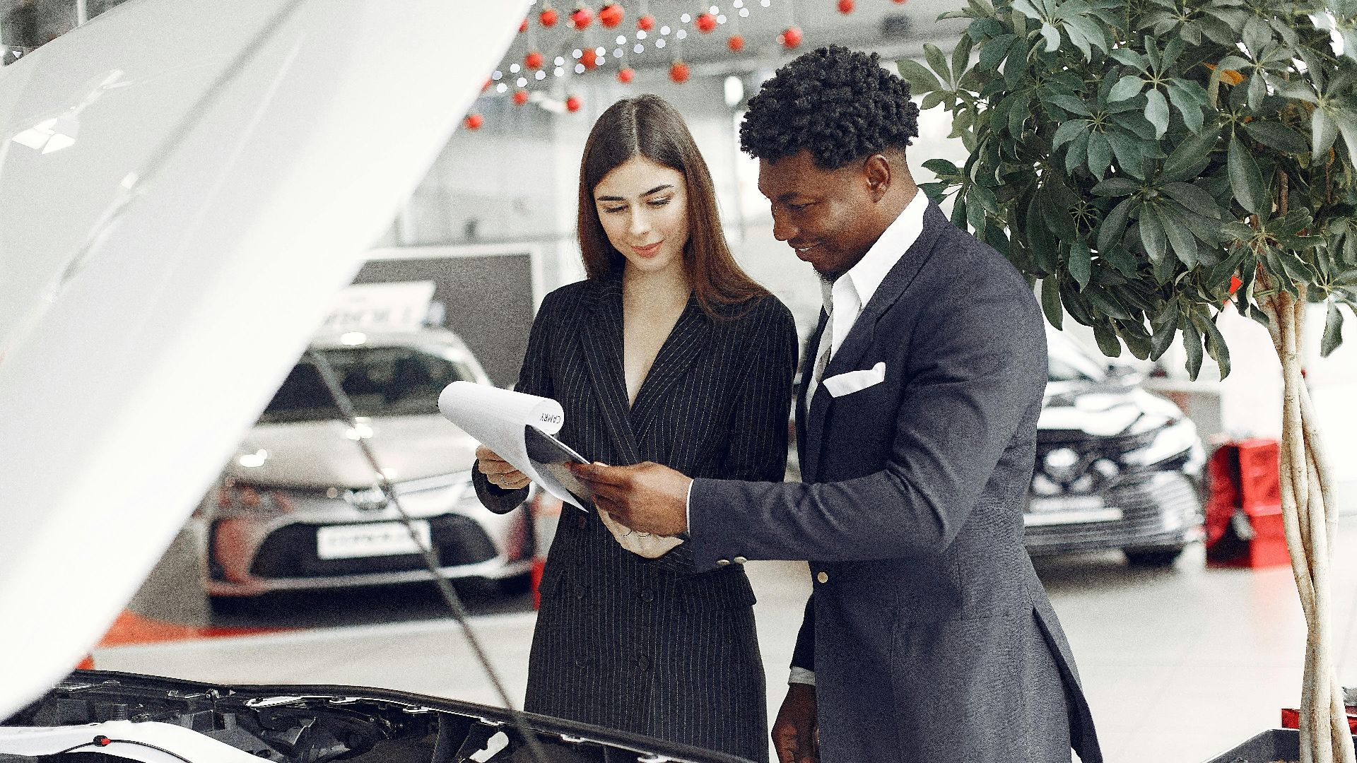 Confident smiling successful black businessman in classy formal suit standing near open motor hood in car showroom and discussing car characteristics with gorgeous female dealer
