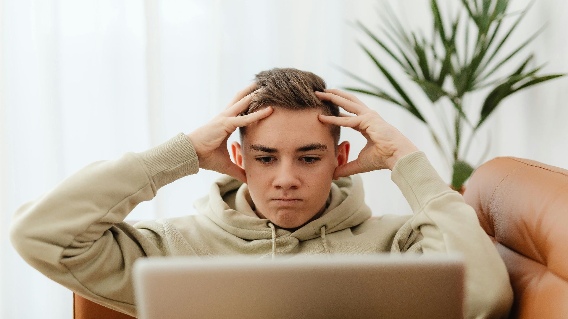 A young man in a hoodie concentrates on his laptop, seated indoors, conveying stress or frustration.