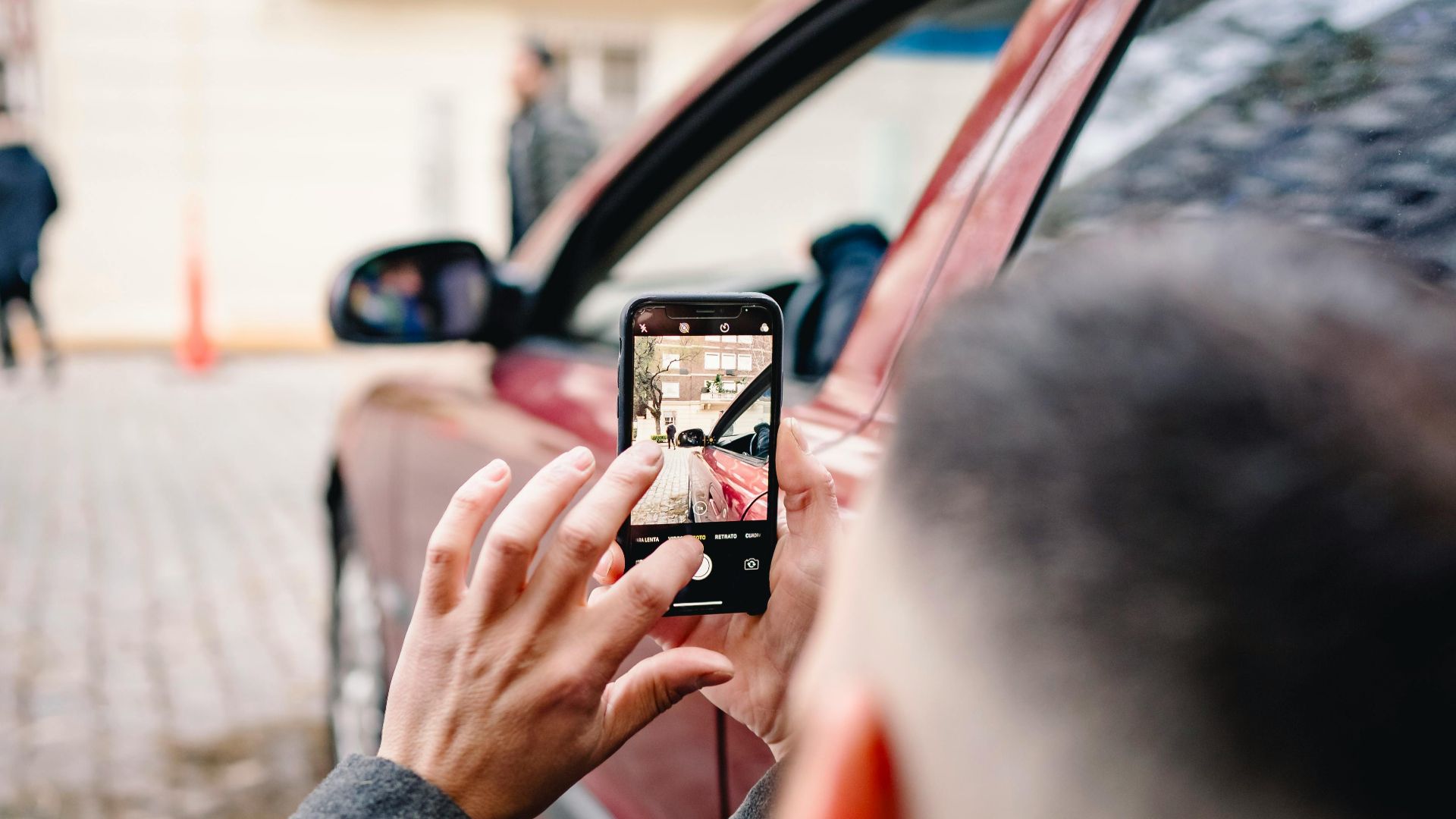Man using smartphone to take pictures of an urban scene beside a red car.