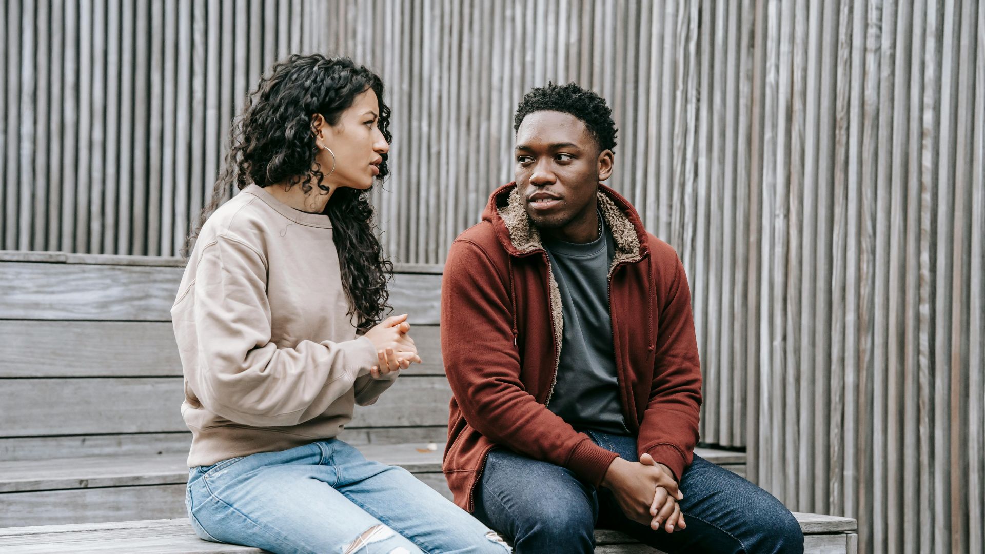 Frustrated ethnic girlfriend and African American boyfriend sitting on wooden step and looking at each other while having dispute on street