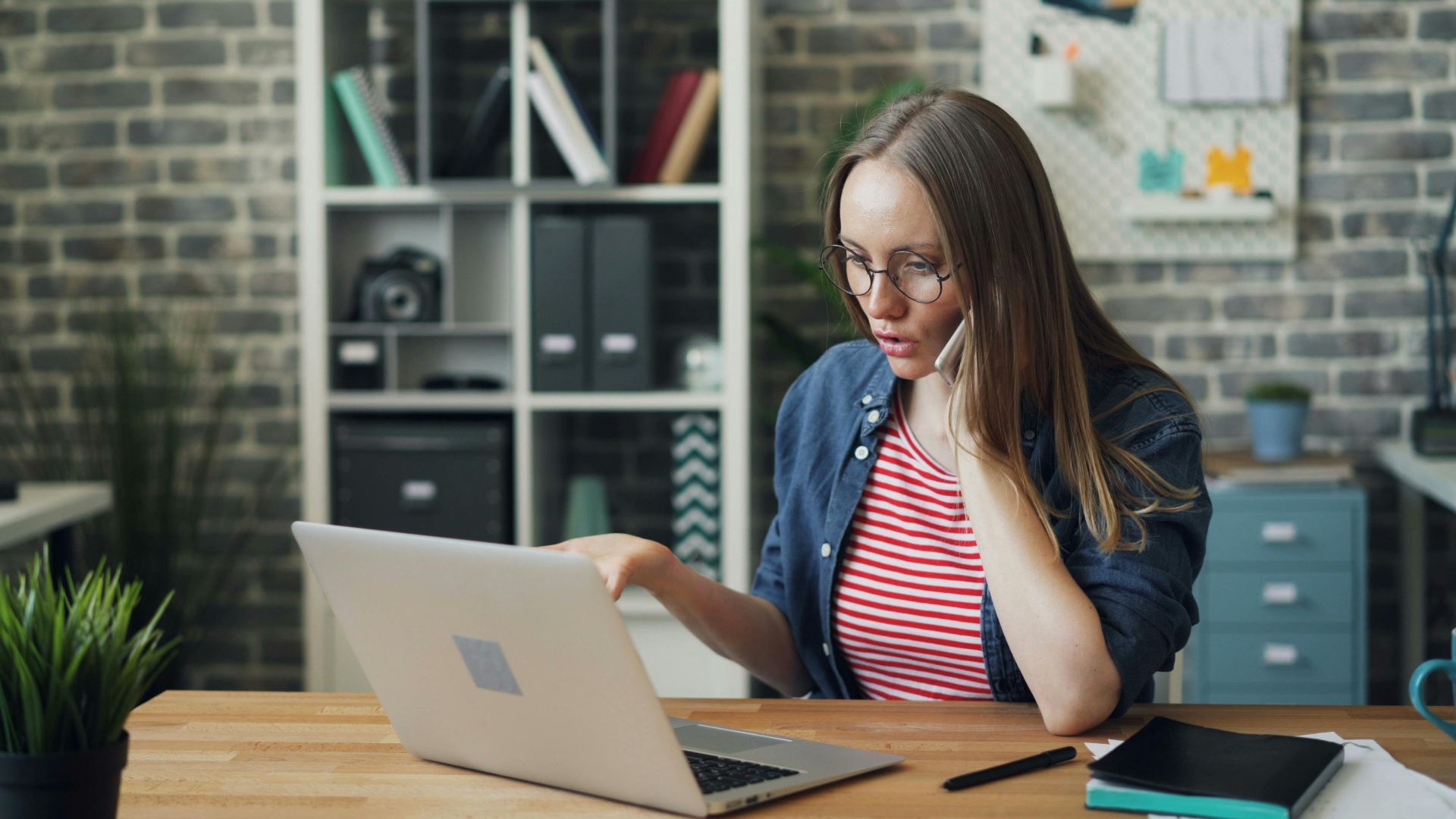 Focused young woman working on laptop while talking on a phone in a stylish modern office.