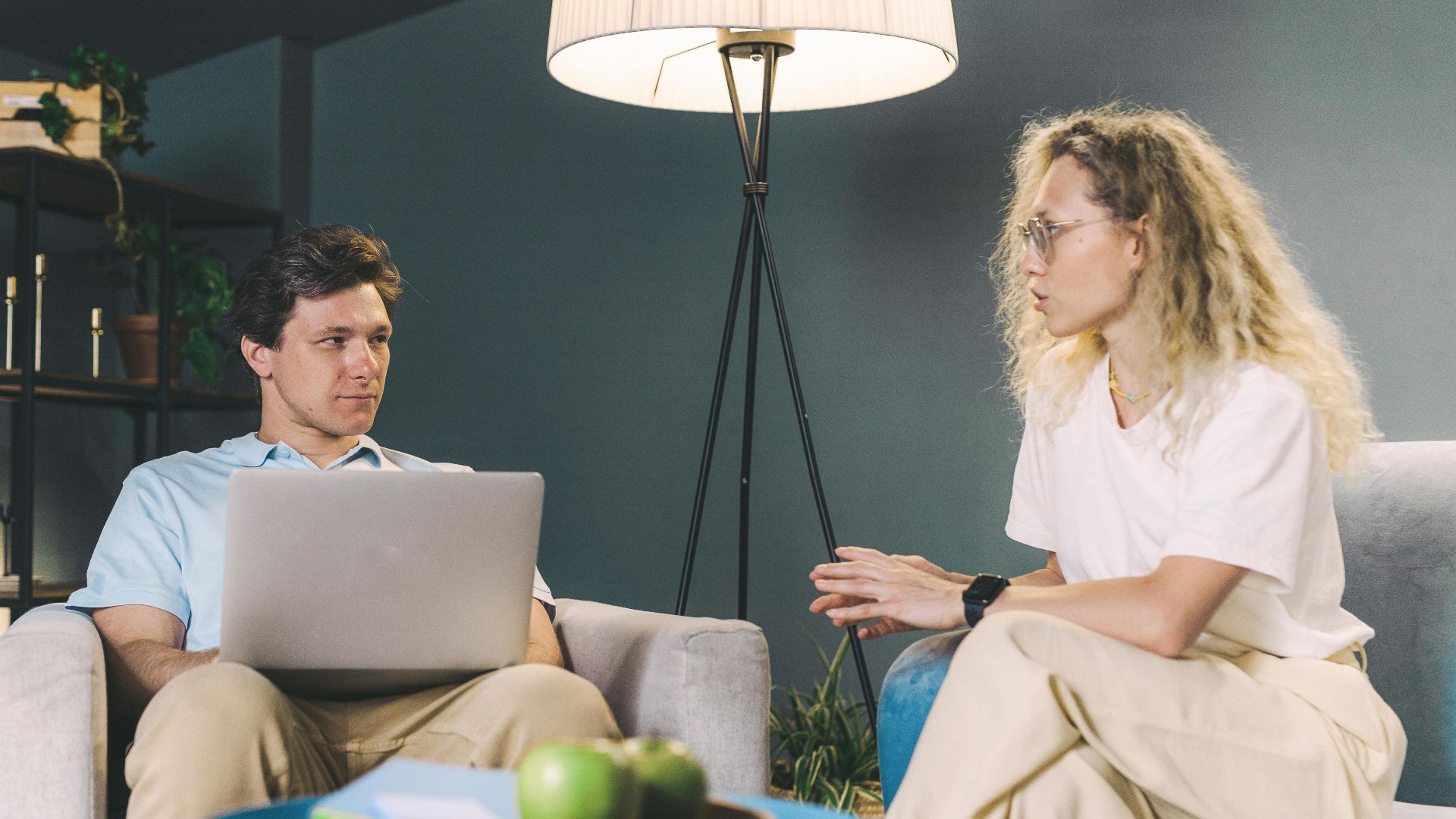 Two adults discuss work while sitting on sofas in a cozy, modern living room setting.