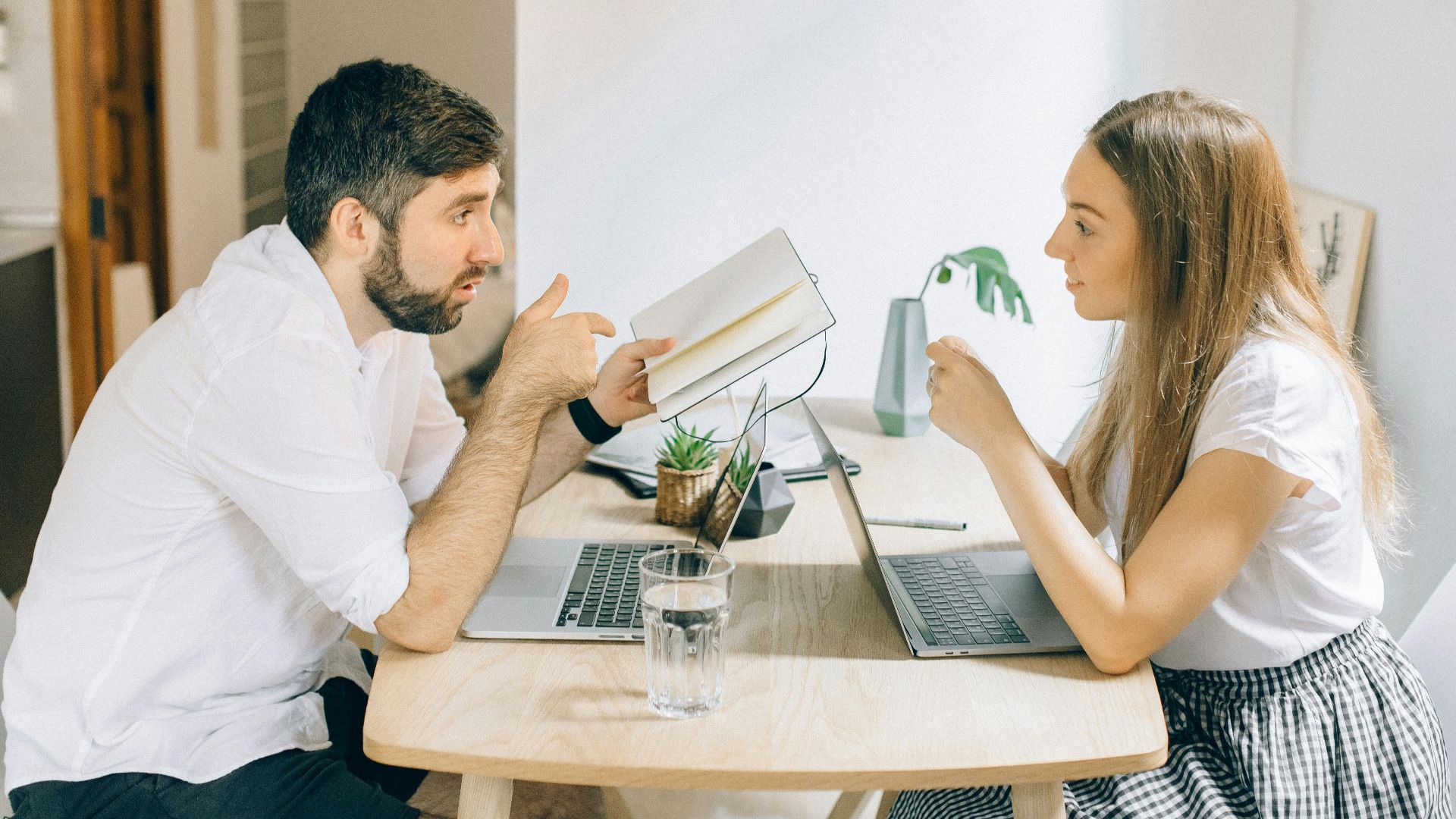 A man and woman working from home on laptops, engaged in discussion at a wooden table.
