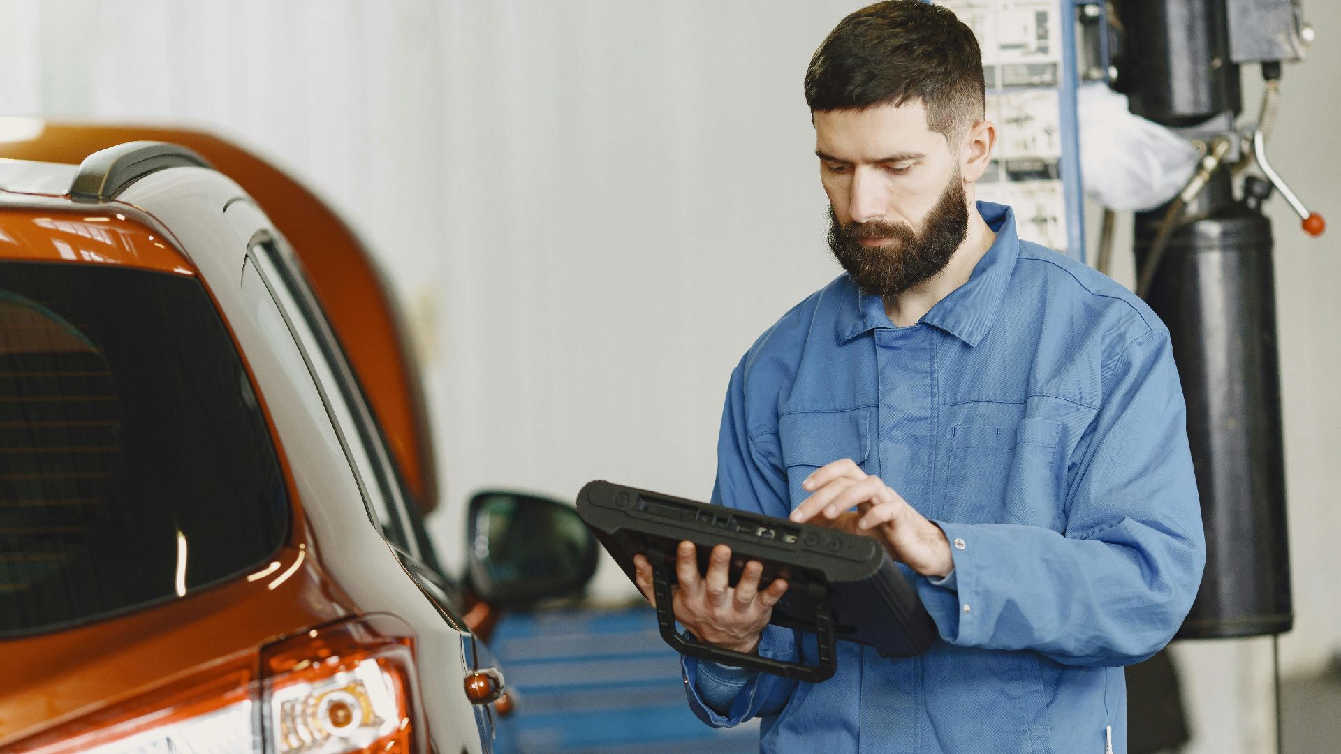 A mechanic in coveralls using a diagnostic tool at an auto repair shop beside a car.