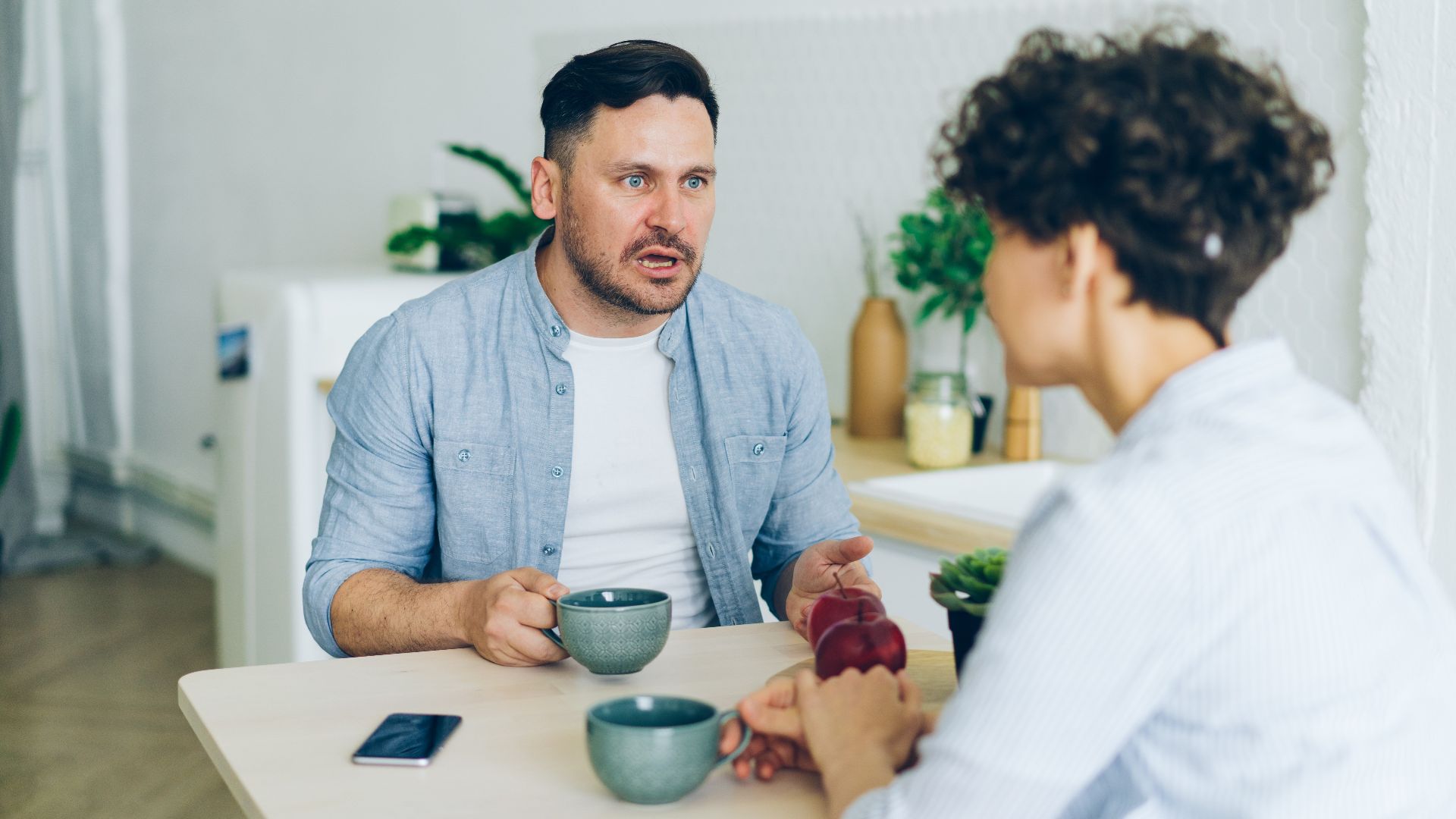 Couple having a serious conversation at the kitchen table with coffee cups.