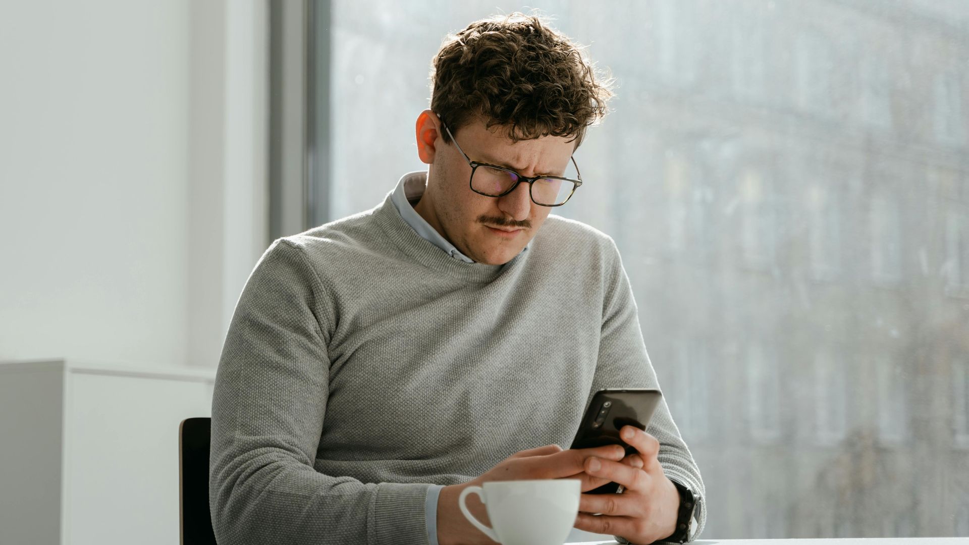 Adult man wearing glasses using smartphone in sunny office space, holding a mug.