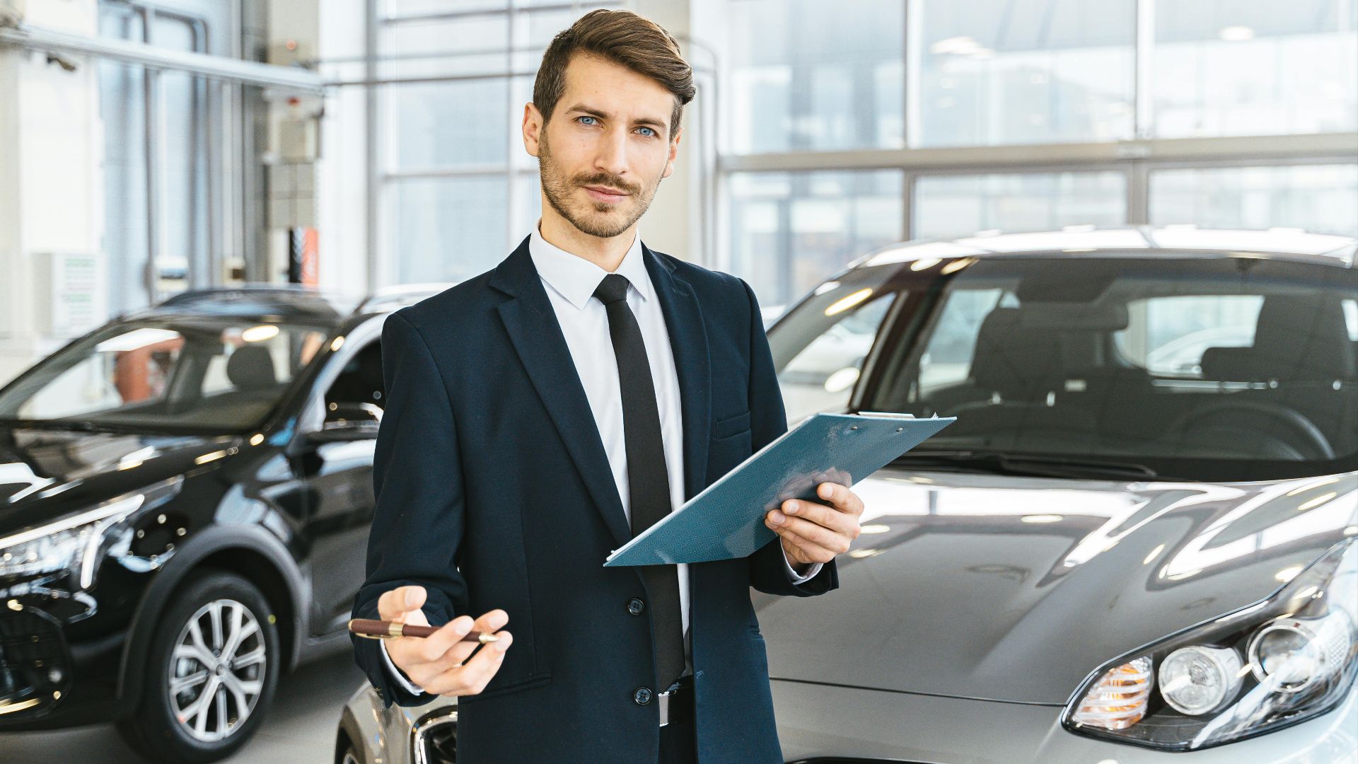 Professional car dealer in business suit holding clipboard in a bright car showroom.