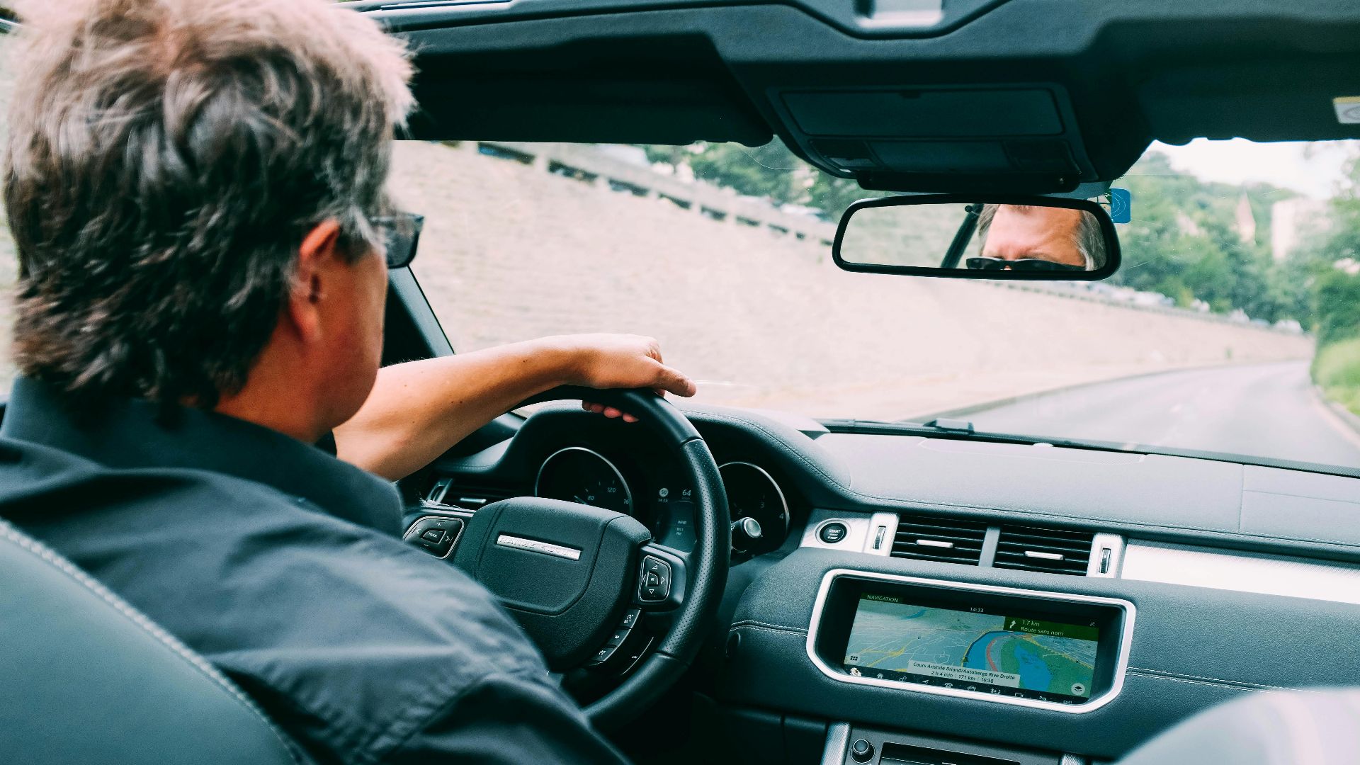 A man driving a convertible car on a scenic highway with a focus on the dashboard.