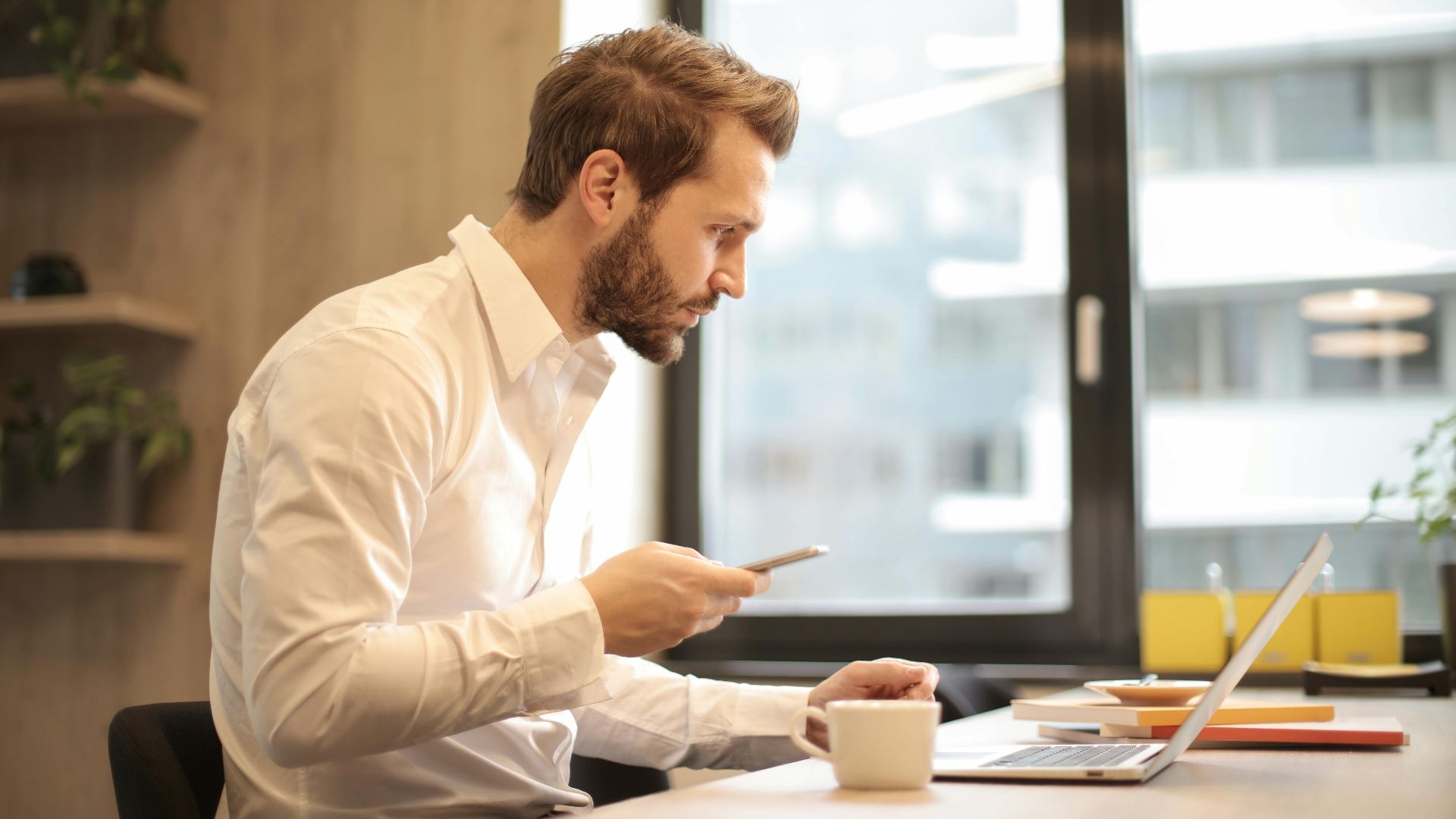 Focused businessman working on laptop while checking smartphone in modern office.
