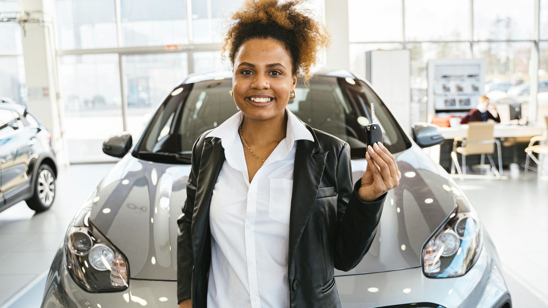 Smiling woman holding car keys in a dealership showroom, celebrating her new car purchase.
