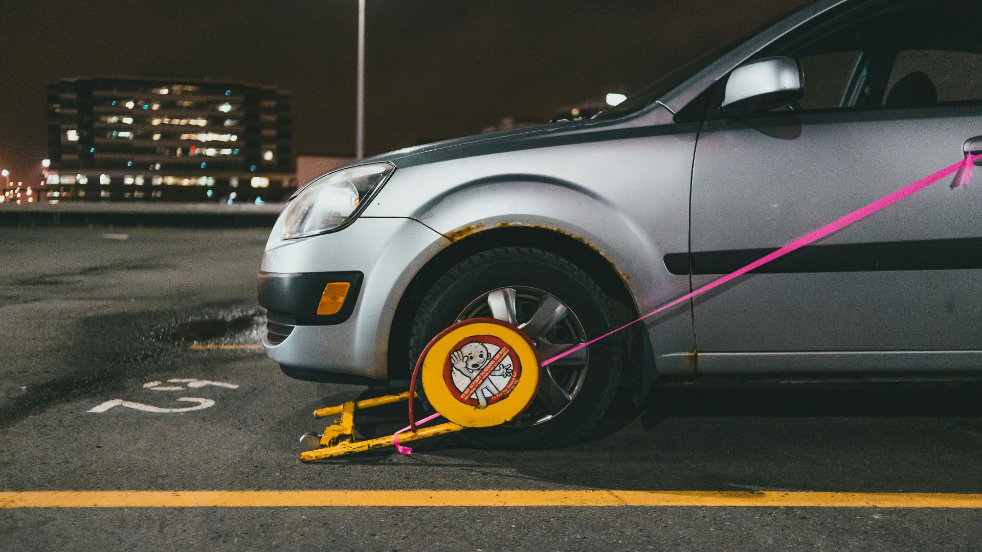 A silver car is immobilized with a wheel clamp in a dimly lit parking lot at night.