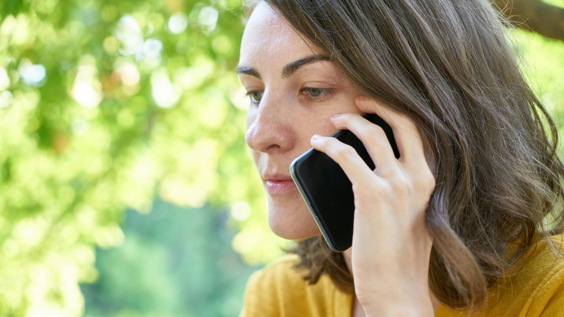 Side profile of a woman using a smartphone outdoors in a sunlit park, wearing a yellow top.