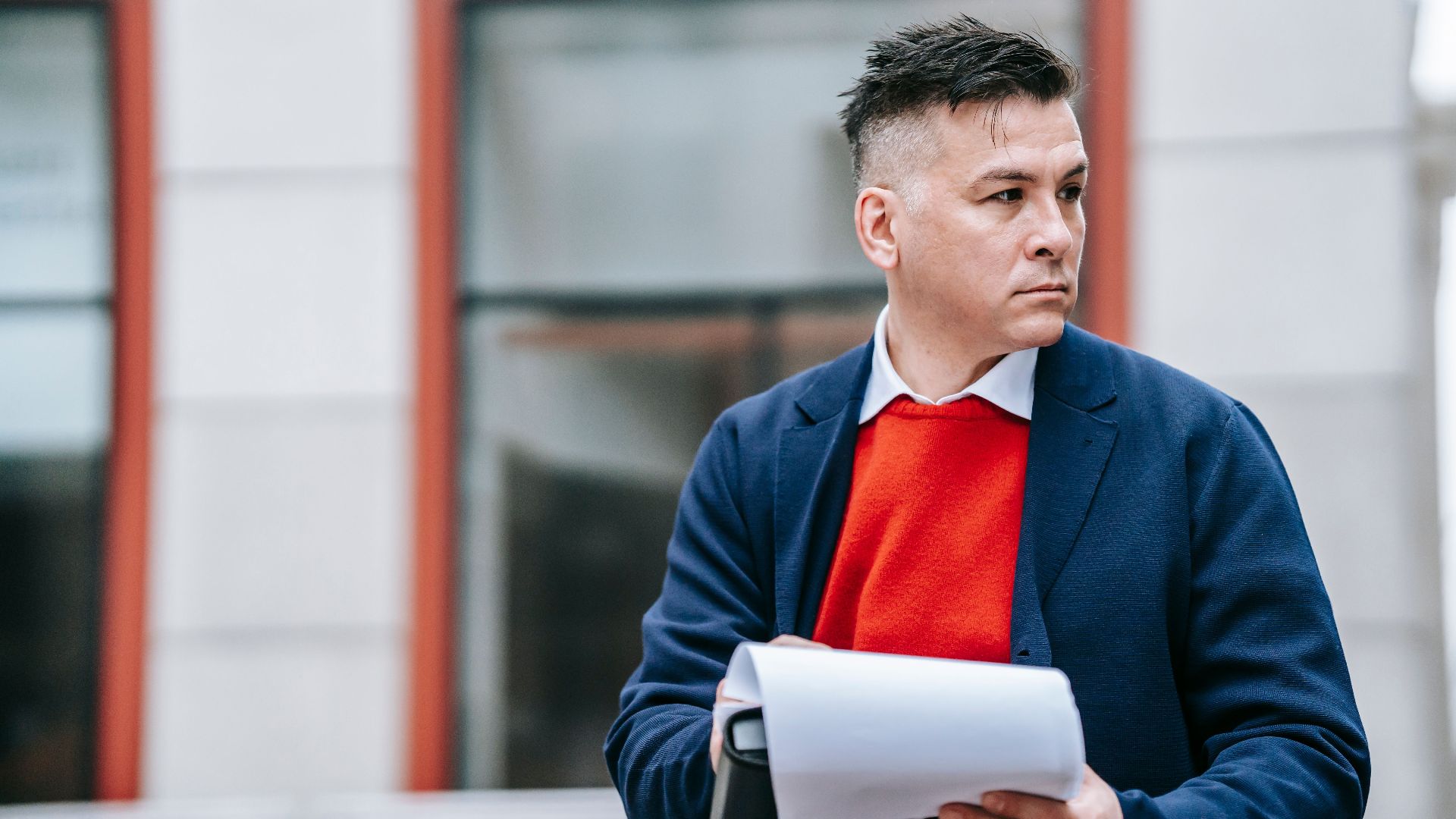 A serious man holding papers, dressed professionally in an urban environment.