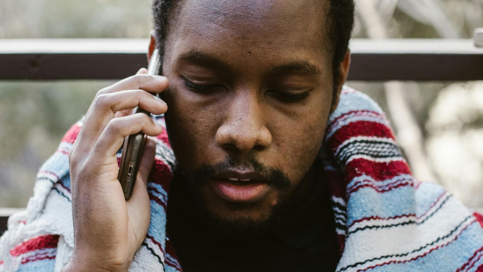 A man wrapped in a striped blanket talks on a phone outside.