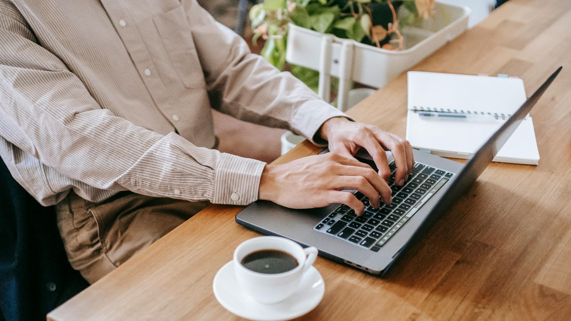 High angle of crop anonymous male freelancer in casual clothes sitting at table and using netbook while drinking cup of coffee and notebook