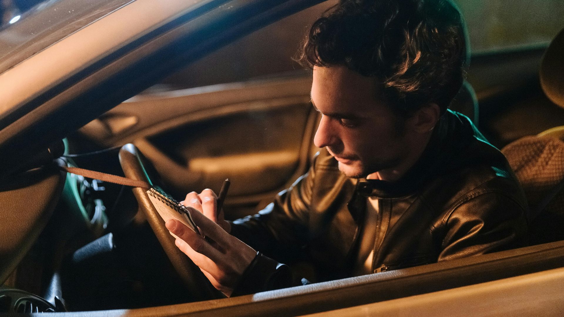 A man sitting in a car at night, writing notes in a notebook.