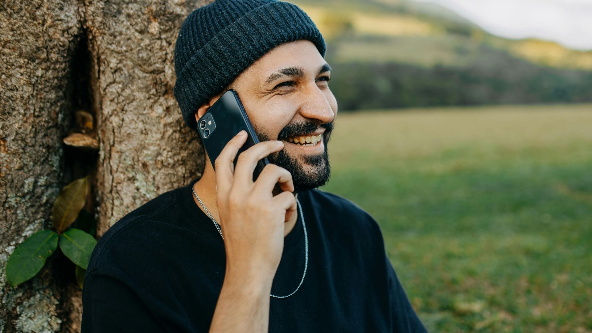 Bearded man in beanie enjoys a phone conversation against a serene rural backdrop.
