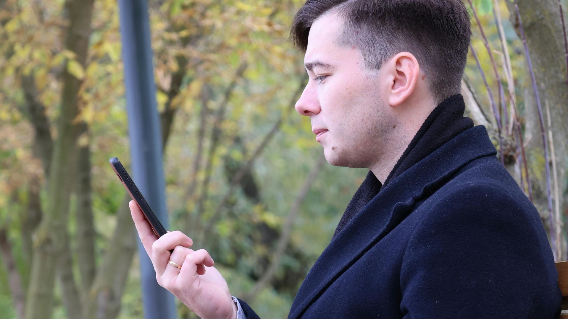 Young man sits on a bench in a fall park, engrossed in his smartphone.