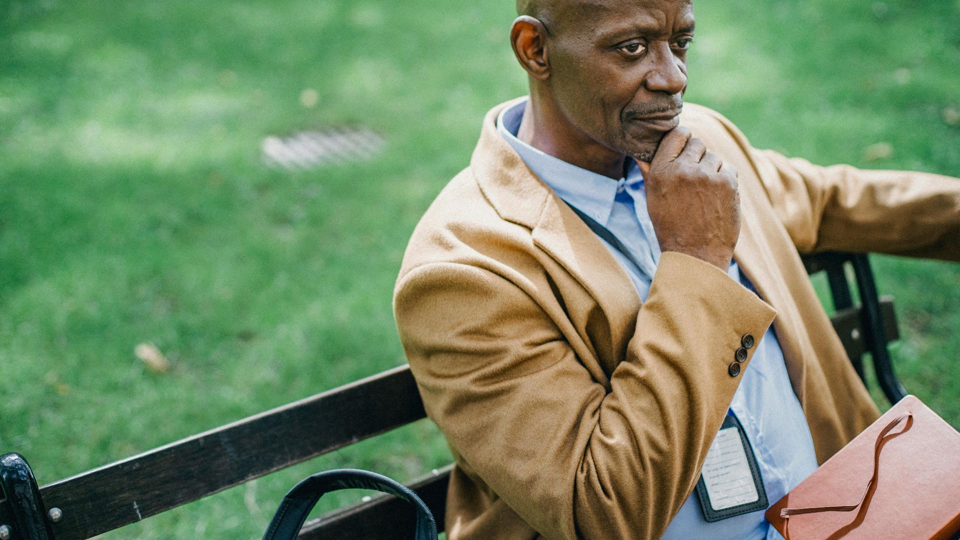 Crop pensive well dressed ethnic male entrepreneur touching chin while resting on bench near lawn and looking away