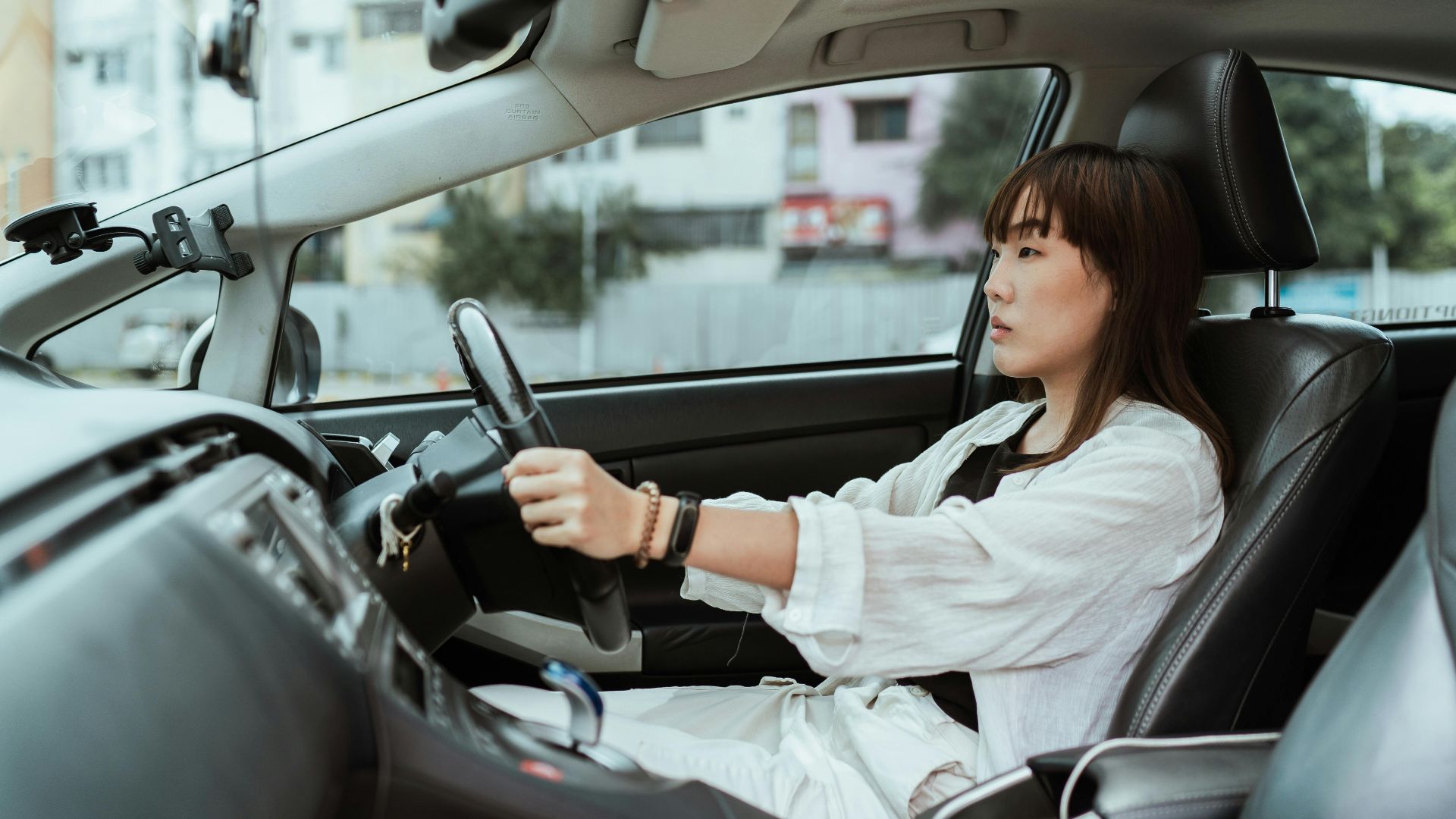 Side view unemotional Asian female in casual white clothes steering right hand drive car along urban streets in daytime