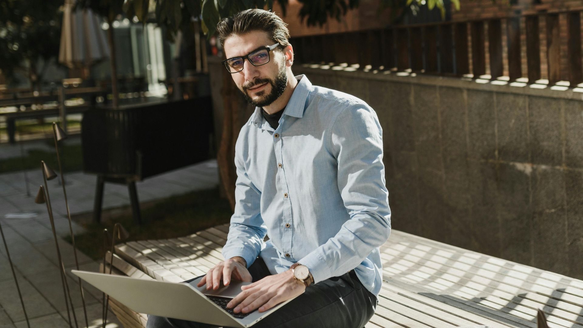 A young caucasian man with eyeglasses working on a laptop outdoors on a sunny day.