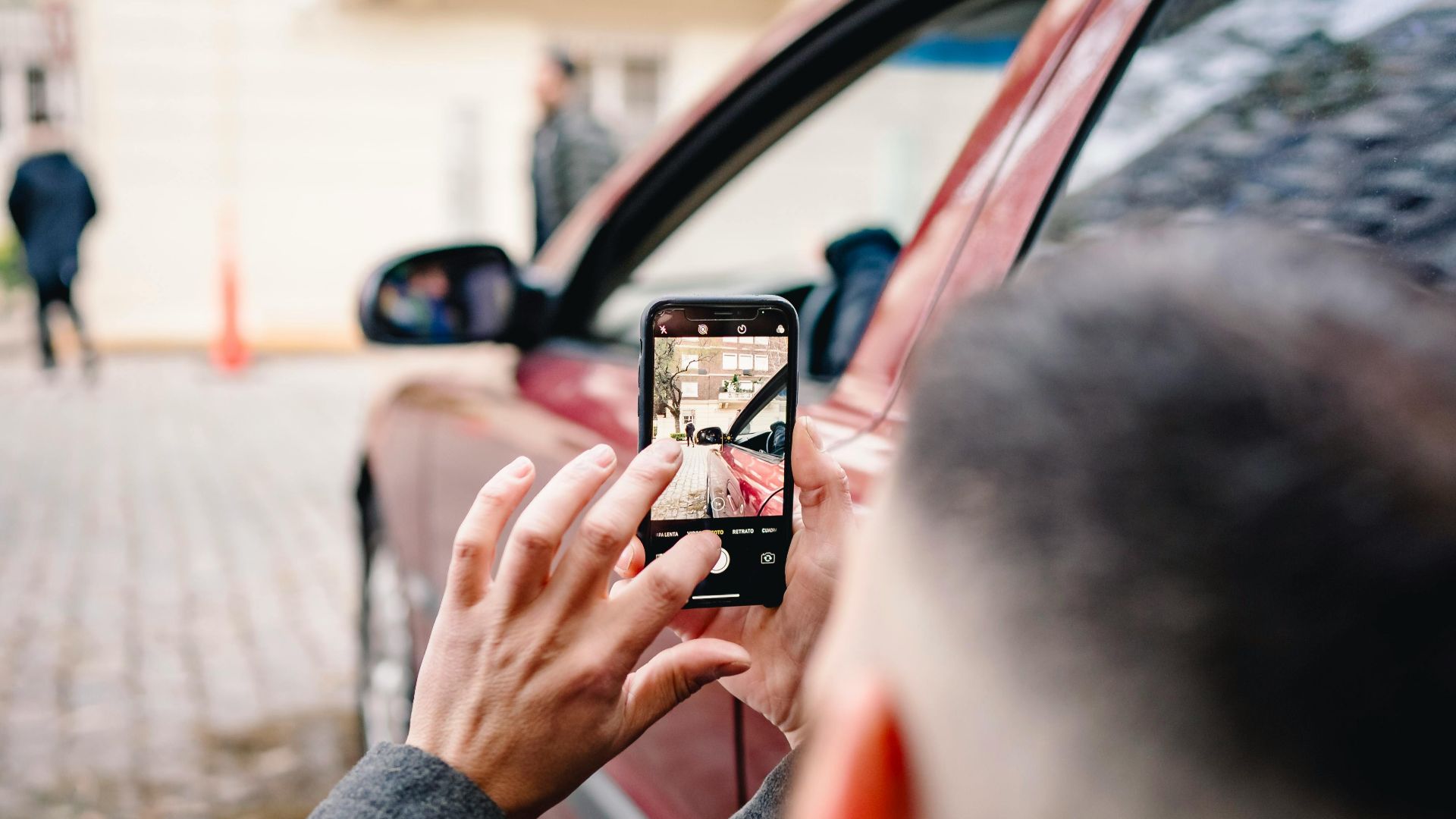 Man using smartphone to take pictures of an urban scene beside a red car.