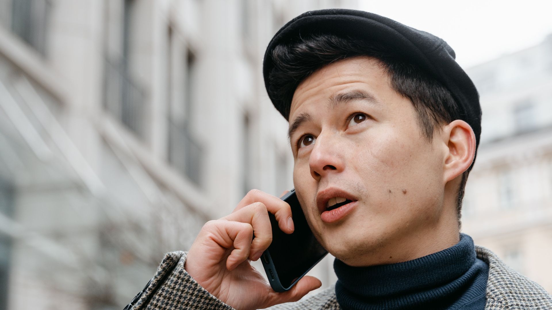 Stylish man in a city talking on a smartphone outdoors, wearing a coat and hat.