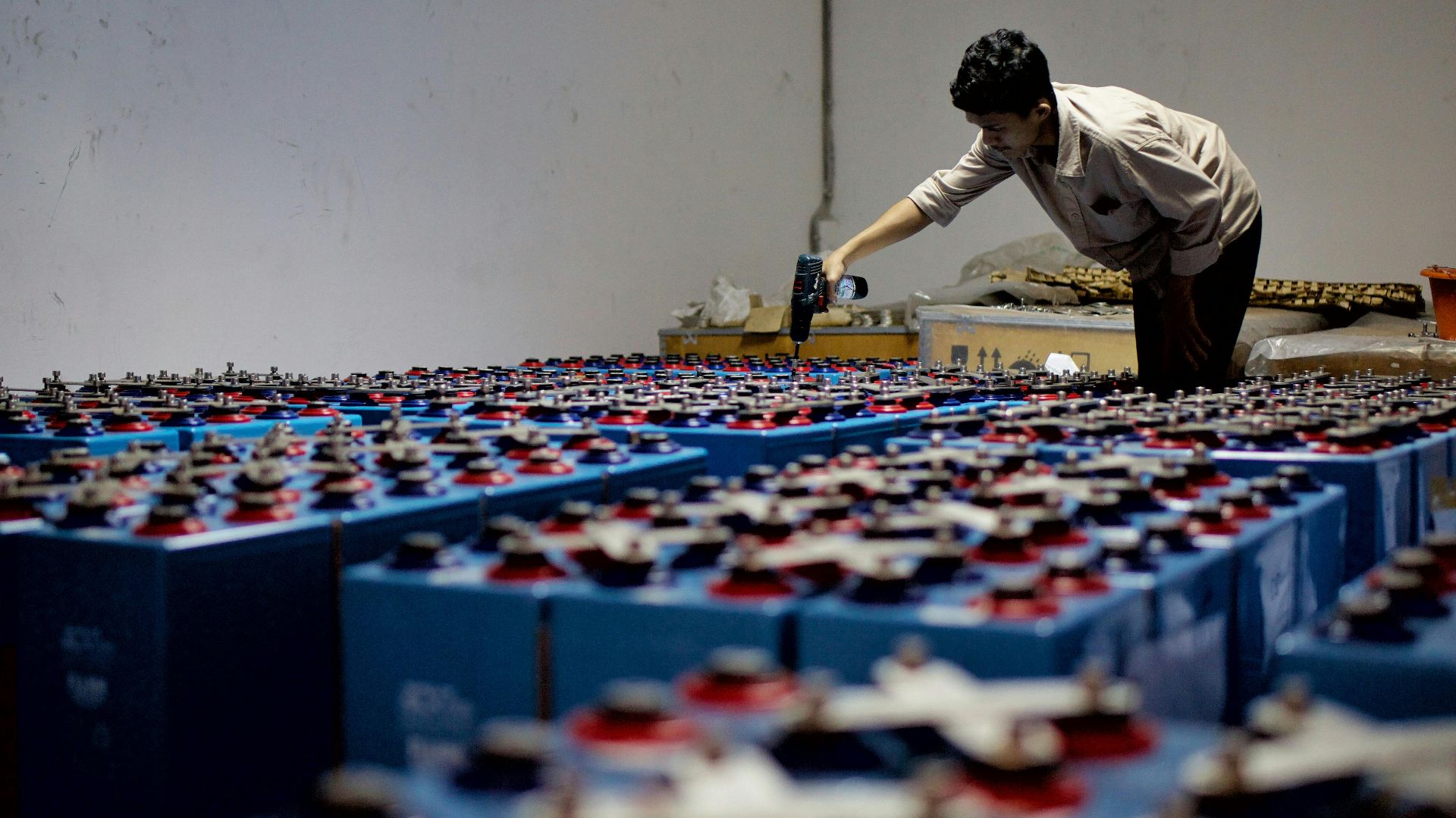 A worker checking many industrial batteries inside a facility. Indoor, industrial setting.