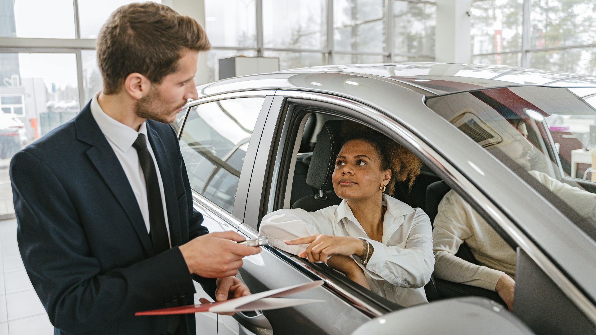 A car dealer hands keys to a woman sitting inside a vehicle in a car showroom.