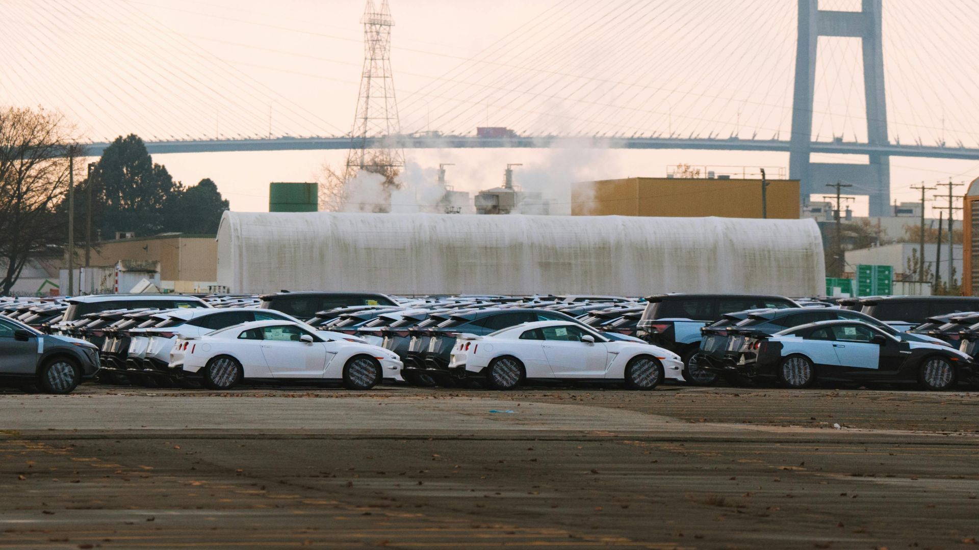 Rows of new cars at a dealership with a bridge in the background under a cloudy day.
