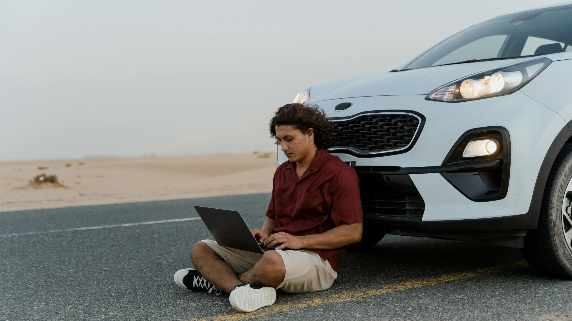 A man using a laptop while sitting on the road beside a parked car in a desert setting.
