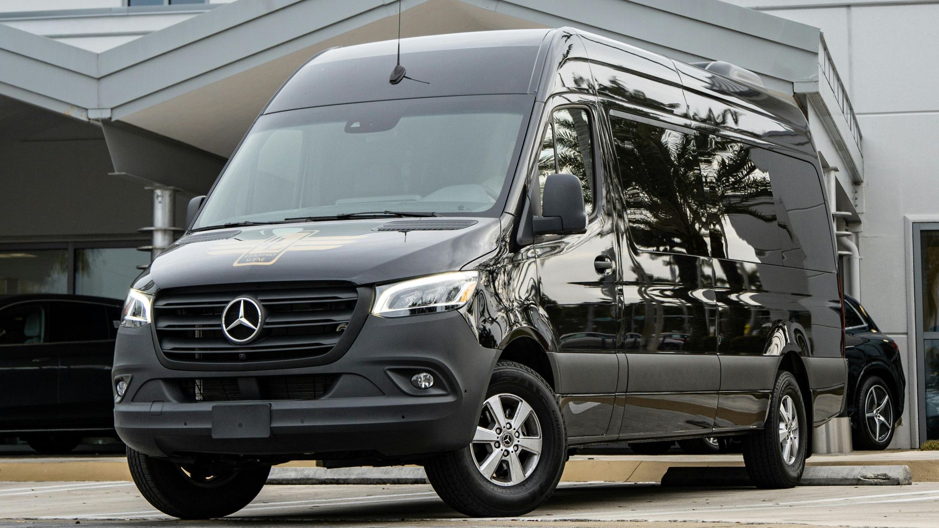 Black Mercedes-Benz Sprinter van parked in front of a modern building under a cloudy sky.