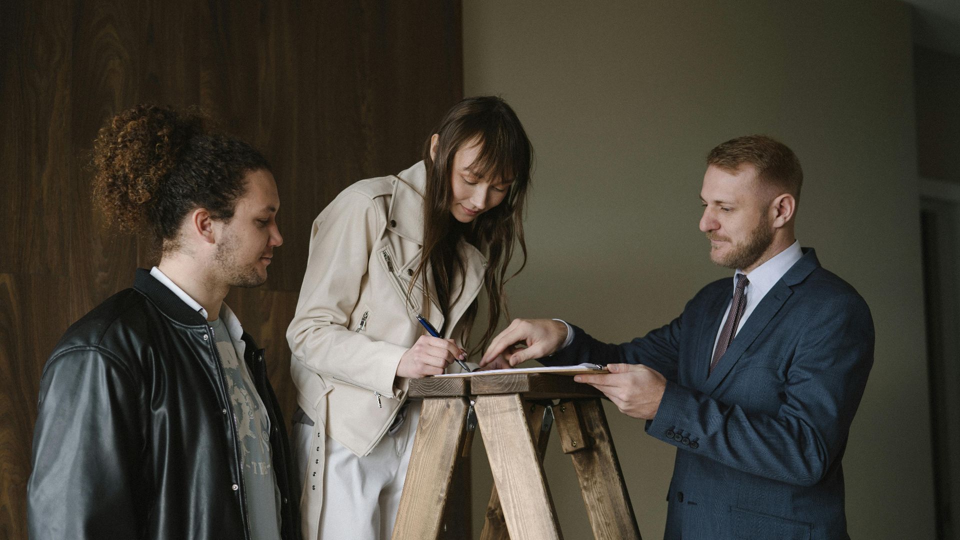 Three individuals signing a contract on a ladder inside a new house, symbolizing a real estate deal.