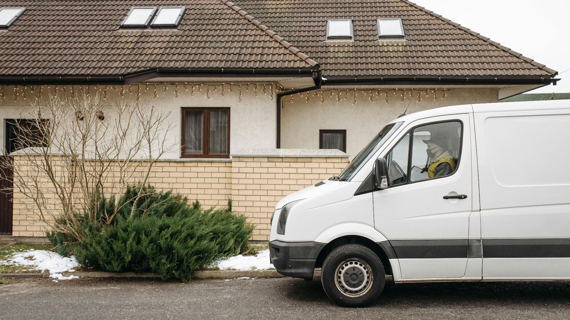 White delivery van in residential area during winter, parked in front of a house with festive lights.