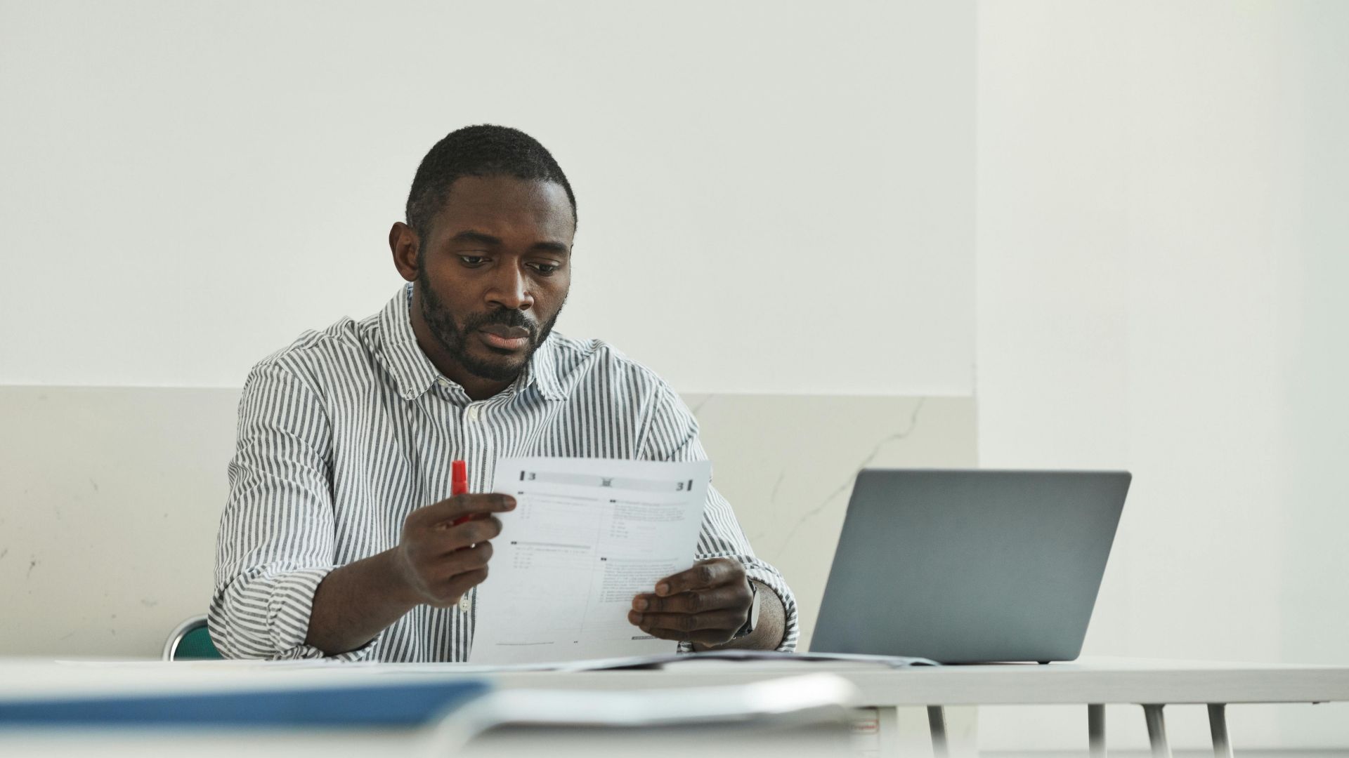 An adult man attentively grading test papers at his workspace with a laptop.